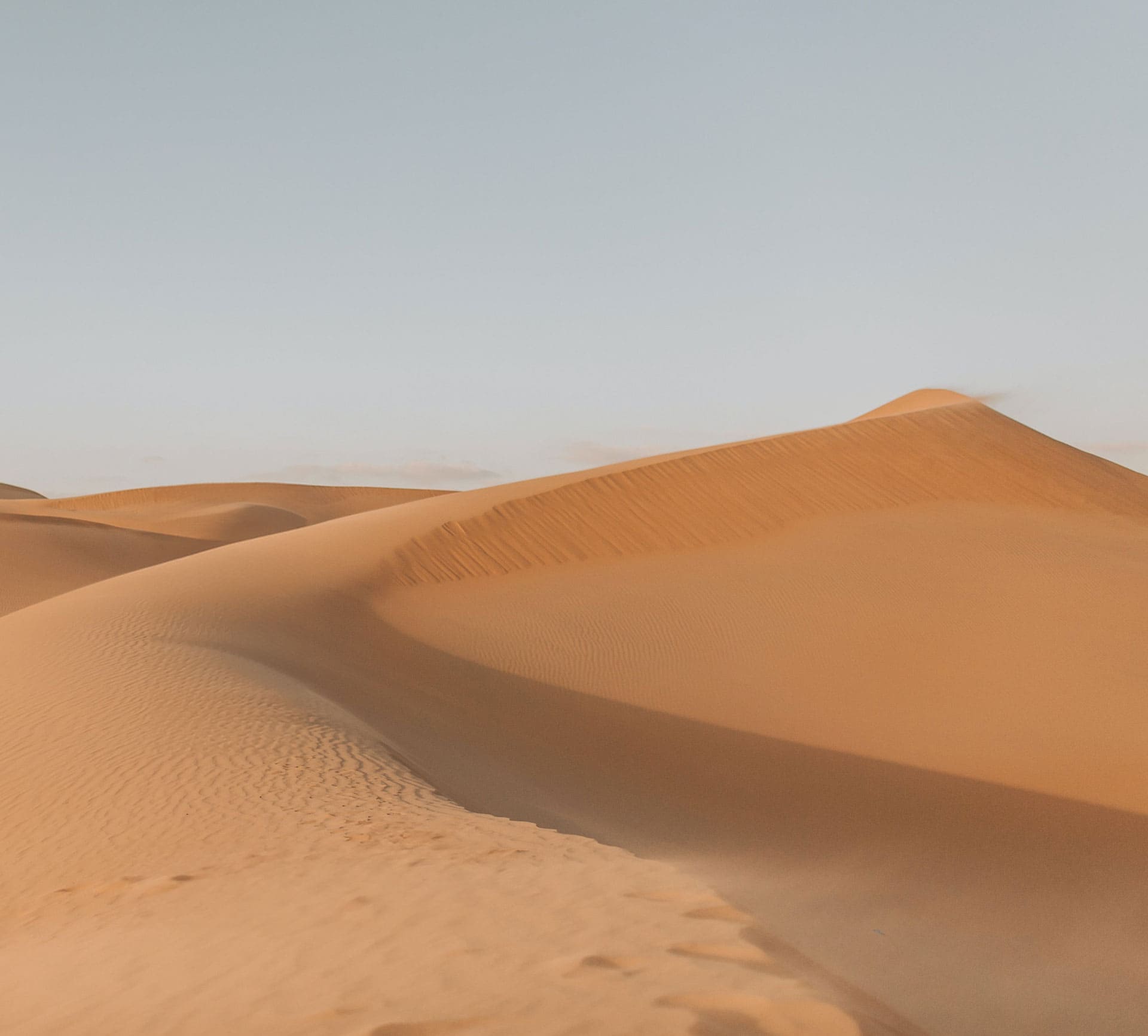 This photograph captures the ethereal beauty of vast desert dunes. The gentle curves and ridges, shaped by the whims of the wind, dance under a soft golden light, offering a mesmerizing symphony of shadow and form. The pristine ambiance is amplified by the expansive sky, creating a harmonious blend of tranquility and grandeur.