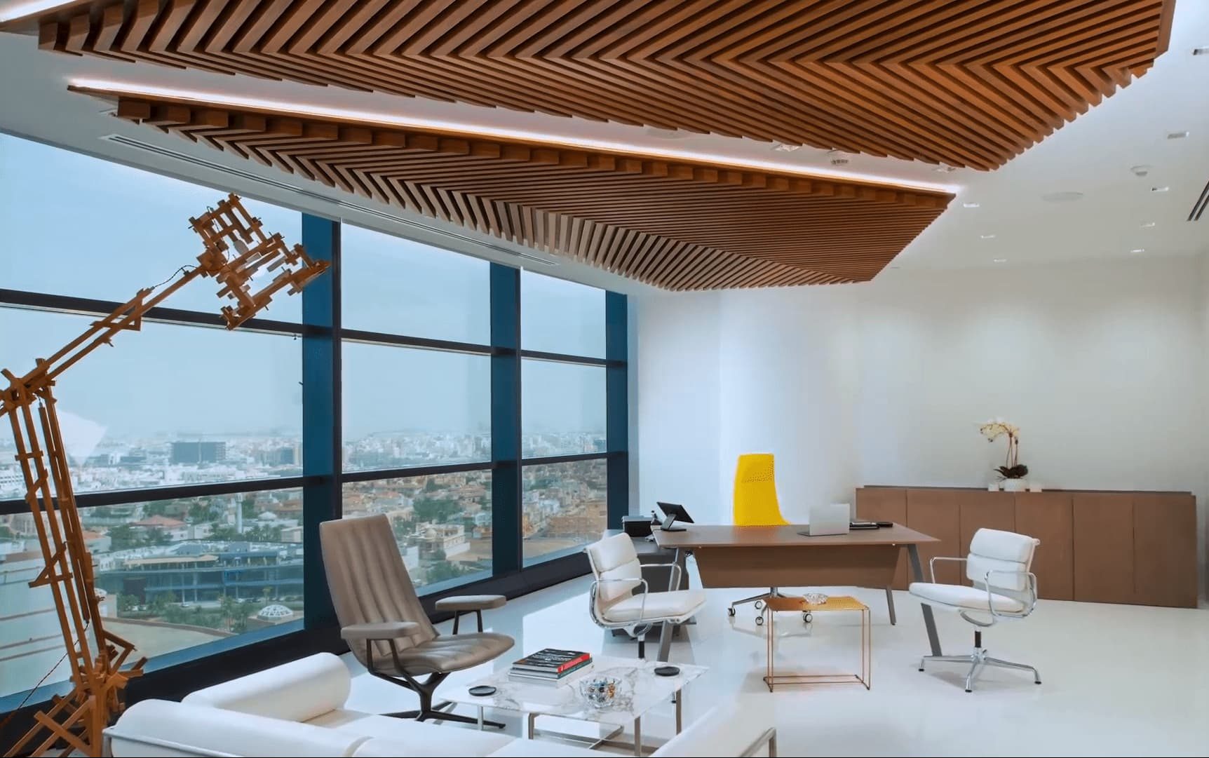 This is an office with a modern aesthetic featuring a sculptural wooden ceiling design, large windows with a city view, a desk with a yellow chair, and a lounging area with a unique wooden sculpture.