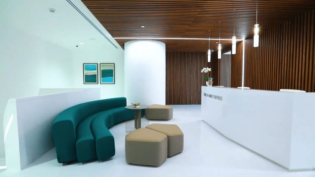 A modern office reception area with a white desk, teal sofa, wooden slats on the ceiling, and pendant lights.