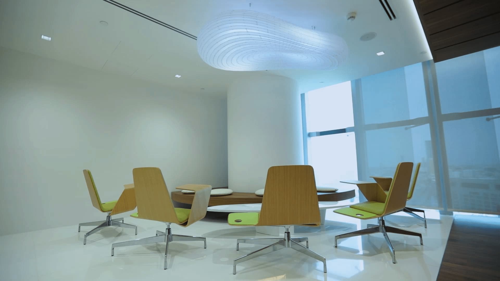 A modern meeting area with green and wood chairs around a small table, under a unique circular light fixture, next to floor-to-ceiling windows.