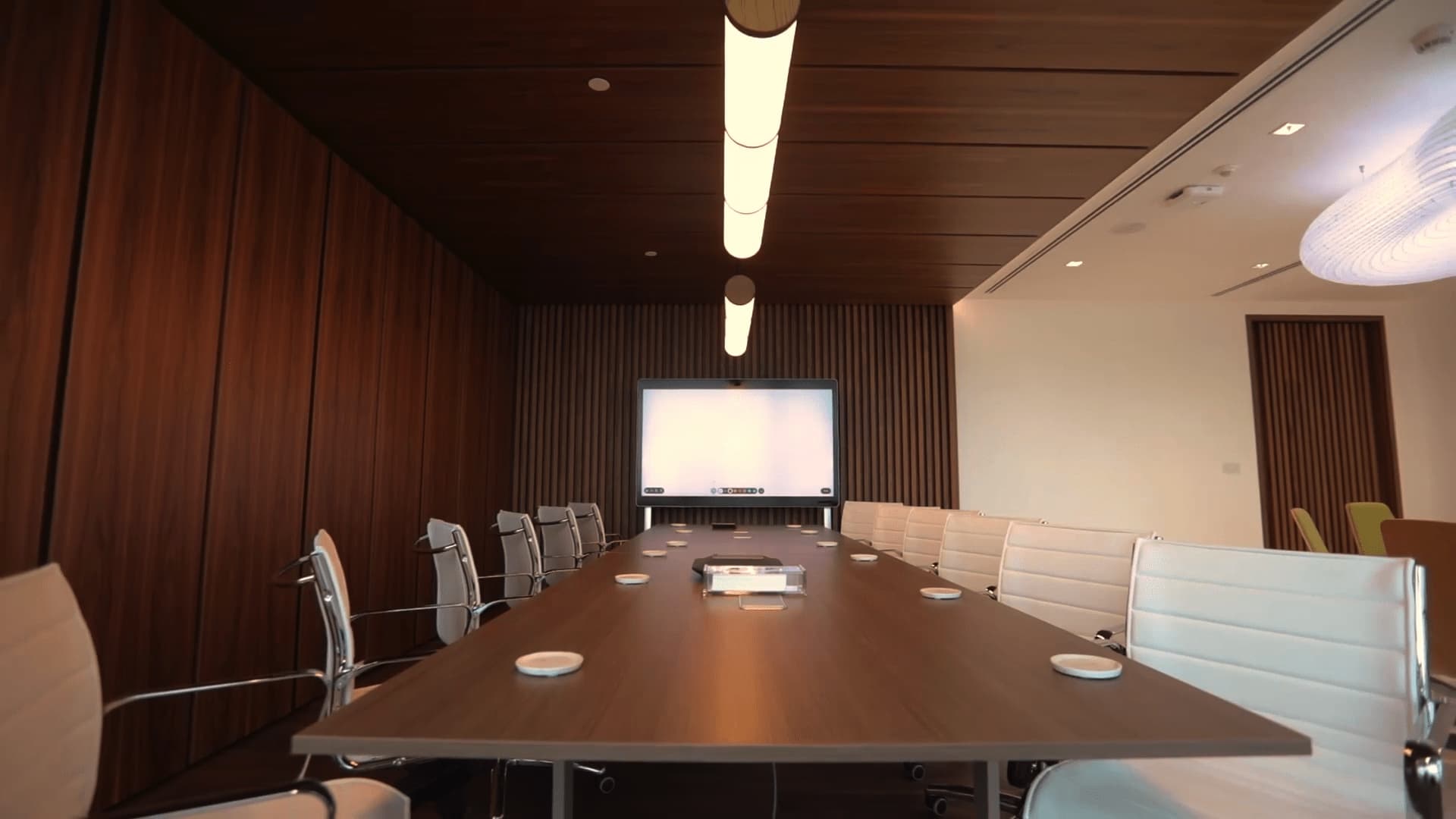 A conference room with a long wooden table, white chairs, a screen for presentations, and a warm lighting scheme complemented by wood-paneled walls.