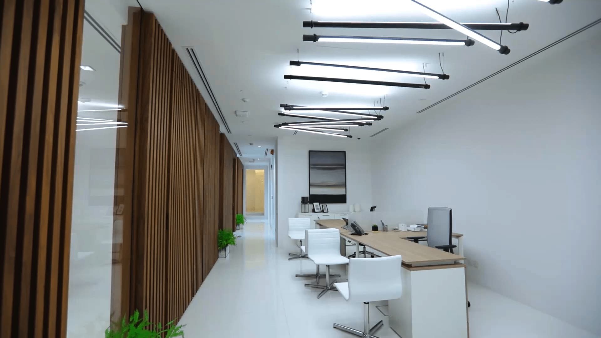 This image displays a modern, minimalist office with a long desk, white chairs, track lighting, wooden slat walls, and indoor plants, creating a bright and airy workspace.