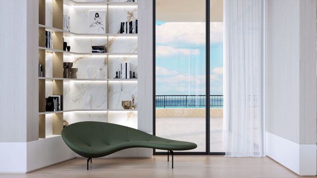 A modern reading nook with a unique green chaise lounge, beside a white bookshelf with marble backing, overlooking a serene ocean view through floor-to-ceiling glass doors draped with sheer curtains.